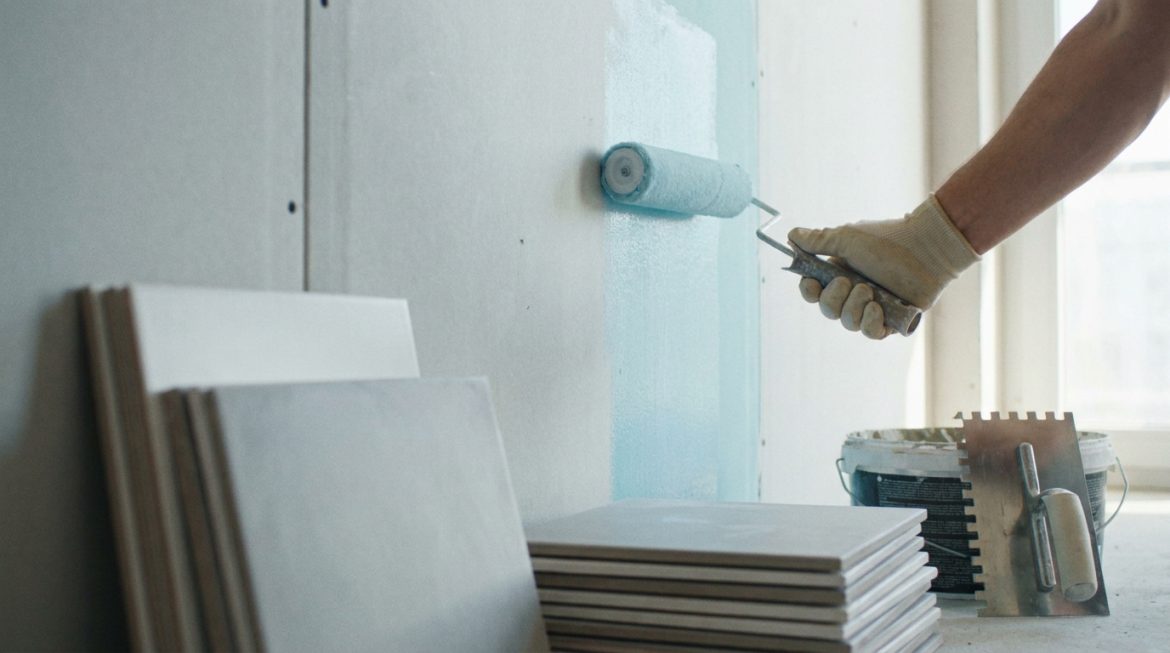 Main gantée appliquant une sous-couche bleue sur une plaque de plâtre avec un rouleau. Des carreaux et outils de pose sont au sol.